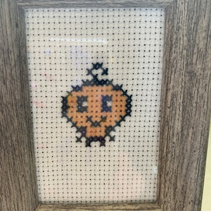 May include: A framed cross-stitch of a small orange pumpkin with a black stem and a black face.