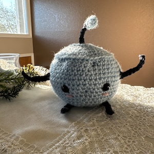 May include: A light blue crocheted plush toy with black yarn arms and a tail. The toy has a round body with a stitched-on face and black eyes.