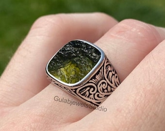 May include: A silver ring featuring a square-cut, textured dark green moldavite stone set in a band with intricate black engraved scrollwork patterns. The text GulabjewelsStudio is visible on the side of the metal band of the ring.