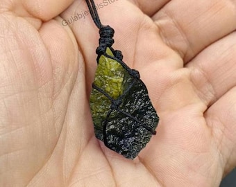 Può includere: Un ciondolo in pietra grezza e testurizzata, nei toni del verde oliva e del verde bosco scuro, fissato in una montatura in cordoncino di macramè nero. La pietra ha una forma irregolare e frastagliata, sospesa a una sottile collana di corda nera.