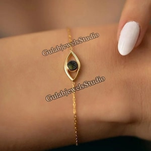 Puede incluir: Un brazalete de cadena dorada con un pequeño dije en forma de ojo con una piedra preciosa verde oscura. El dije tiene la forma de un ojo con un fino contorno dorado.