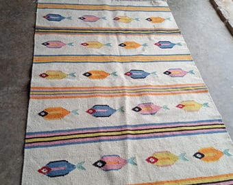 May include: A rectangular, off-white rug with a repeating pattern of colorful fish. The fish are in shades of orange, yellow, pink, and blue, with black eye details. Horizontal stripes in various colors separate the fish rows. The rug has a fringed edge.