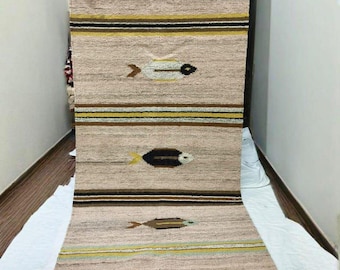 May include: A beige and brown woven rug with a fish pattern. The rug features horizontal stripes in brown and yellow, with fish motifs in various colors. The rug is rectangular and appears to be handmade.