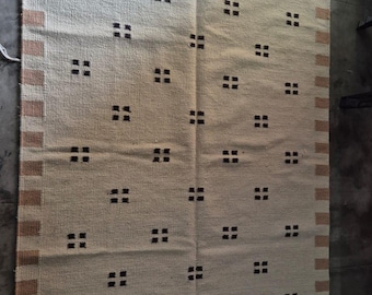 May include: A rectangular woven rug featuring a cream-colored base with a repeating pattern of small dark brown square motifs. The rug is bordered by wide tan and cream stripes on the left and right sides, finished with white fringe at both ends.