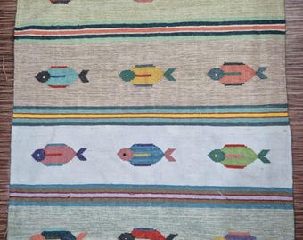May include: A rectangular woven rug featuring horizontal stripes in shades of green, beige, white, and gray. The rug is decorated with a repeating pattern of small, multi-colored fish motifs in red, blue, yellow, and green, finished with white fringe.