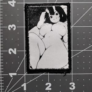 May include: Black and white embroidered patch featuring a cartoon illustration of a female figure with horns. The figure is reclining, with a detailed outline and shading. The patch has a black border.