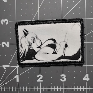 May include: A rectangular patch with a black border features a black and white anime-style illustration of a person lying down. The person has long hair, and is wearing a striped top. The patch is on a gray surface with measurement markings.