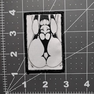 May include: A rectangular patch with a black border features a black and white illustration of a stylized figure. The figure has a large, rounded bottom and a small head with large eyes. The design is on a white background.