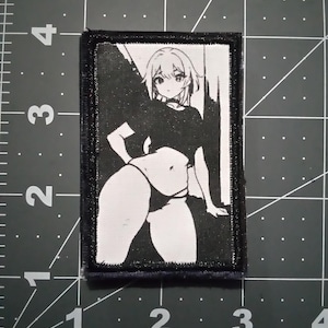 May include: Black and white embroidered patch featuring an anime-style illustration of a woman with long hair wearing a black top and white underwear.