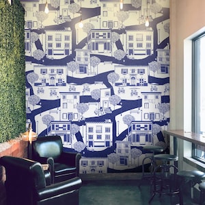 May include: A white wall with a blue and white patterned wallpaper featuring a repeating design of buildings, trees, and streets. The wallpaper is a stylized illustration of a city scene. The wallpaper is in a room with a black leather armchair and a black metal stool.