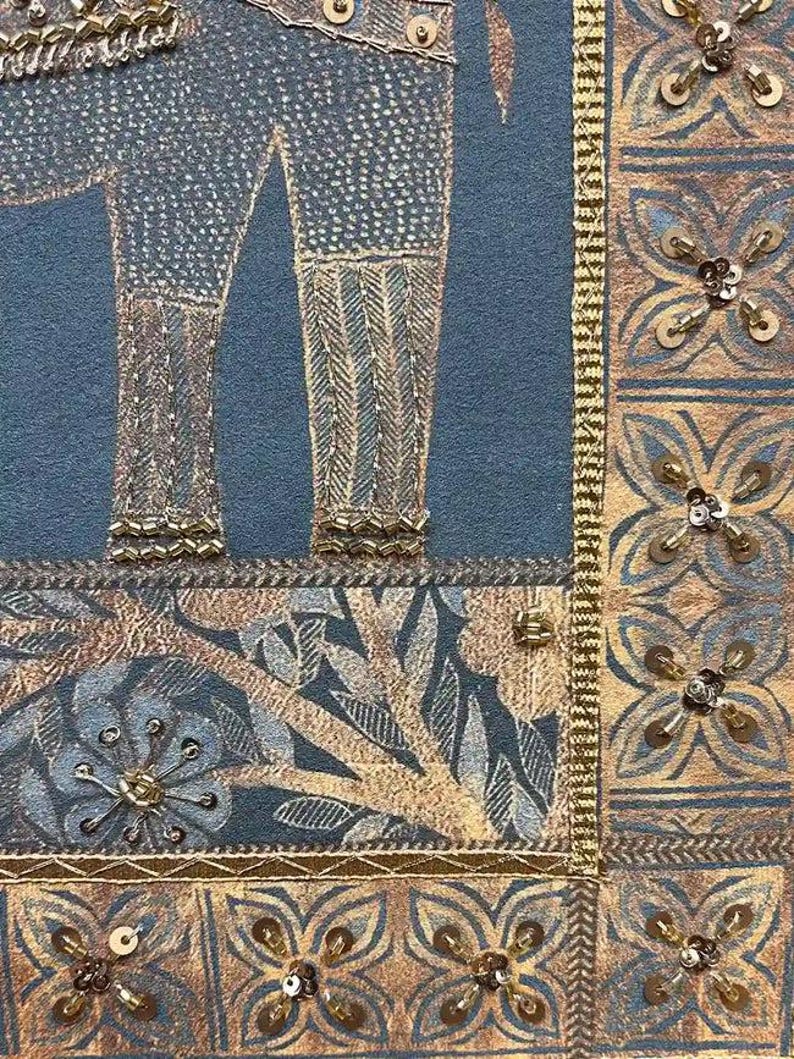 May include: Close-up of a textile with an elephant design. The elephant's legs are detailed with gold thread and sequins against a blue background. The border features floral patterns and gold embellishments, creating a rich, textured look.