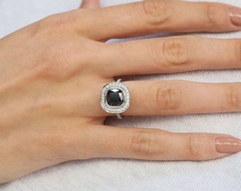 Pode incluir: Um anel de prata com um grande diamante preto quadrado cercado por uma halo de diamantes menores.