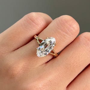 Op de afbeelding: Een gouden ring met een grote, ovaal geslepen diamant. De diamant is gezet met vier klauwen en heeft een heldere, heldere uitstraling. De band van de ring is een delicate goudkleur, en het ontwerp is eenvoudig maar elegant.