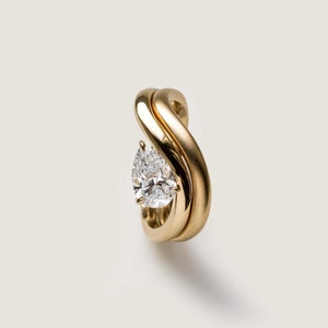 Puede incluir: Un anillo de oro con un diamante de forma de pera engastado en el centro. El anillo está hecho de dos bandas que están entrelazadas.