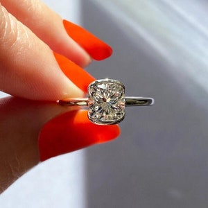 Pode incluir: Um anel de prata com um grande diamante claro de forma quadrada. O diamante está engastado em um engaste de bezel e tem uma forma ligeiramente curva.