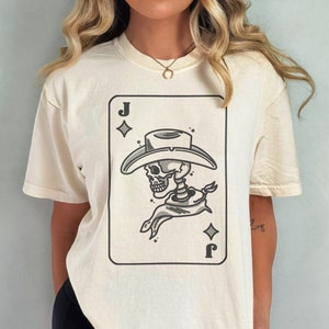 Puede incluir: Una camiseta blanca con un gr&aacute;fico de un esqueleto con un sombrero de vaquero y un pa&ntilde;uelo. El gr&aacute;fico tiene la forma de una carta de juego con la letra 'J' y un s&iacute;mbolo de diamante. La camiseta est&aacute; etiquetada con "COMFORT COLORS" e "IVORY".