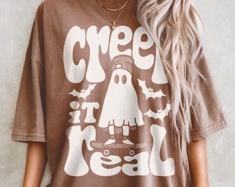 May include: Brown t-shirt with the text "Creep it Real" in white, featuring a ghost on a skateboard and bat graphics. The shirt has a relaxed fit and short sleeves, perfect for Halloween.