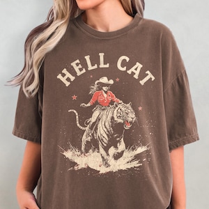 May include: An oversized brown t-shirt featuring a graphic print of a woman riding a tiger. The text "HELL CAT" is printed above the graphic.