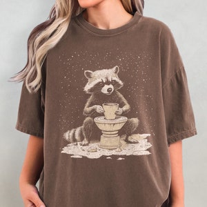 Puede incluir: Una camiseta marrón con un gráfico de un mapache sosteniendo una taza de café en un torno de alfarería. El mapache está sentado en una plataforma con un patrón de estrella.
