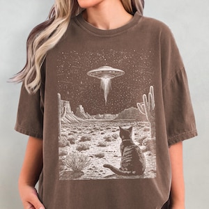 Pode incluir: Uma camiseta marrom com uma estampa gráfica de um gato sentado em uma paisagem desértica com um OVNI no céu. O gráfico é em preto e branco.