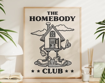 May include: A framed print with a black and white illustration of a house with legs and arms. The house is wearing shoes and giving a thumbs up. The text "THE HOMEBODY CLUB" is printed in black letters on a white background.