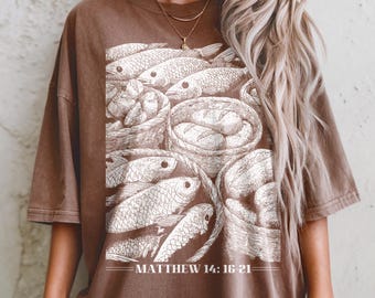 Puede incluir: Camiseta marrón con una ilustración blanca de peces y cestas de pan. El texto "MATEO 14: 16-21" está debajo. La camiseta tiene un corte holgado y mangas cortas.