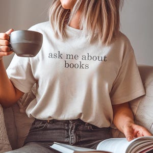 Könnte beinhalten: Hellbeige T-Shirt mit dem schwarzen Text "ask me about books". Die Person hält eine graue Tasse und ein Buch mit weißen Seiten und einem roten Rücken. Die Person trägt eine graue Jeans.
