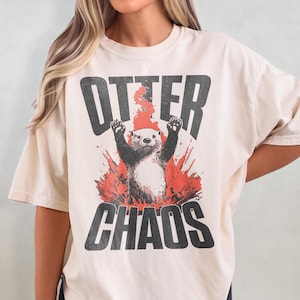 Puede incluir: Una camiseta de color crema con un gr&aacute;fico de una nutria en una explosi&oacute;n roja. El texto "Otter Chaos" est&aacute; impreso en negro en la camiseta.