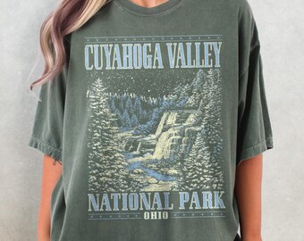 May include: Olive green t-shirt featuring a graphic of Cuyahoga Valley National Park in Ohio. The design includes a waterfall, trees, and the text "CUYAHOGA VALLEY NATIONAL PARK OHIO" in a vintage style. The shirt has short sleeves and a crew neck.