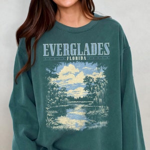 May include: A teal green sweatshirt with a vintage-style graphic print of a Florida Everglades landscape. The text "EVERGLADES FLORIDA" is printed above the image.