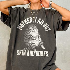 May include: Dark grey t-shirt with the text "MOTHER, I AM BUT SKIN AND BONES" in white. The graphic features a detailed illustration of a seated cat. The shirt has short sleeves and a relaxed fit.