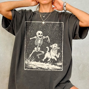 May include: Dark gray oversized t-shirt featuring a black and white graphic of a skeleton running with a dog. The graphic is set within a rectangular frame. The shirt has short sleeves and a relaxed fit.