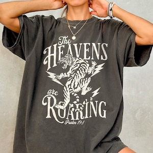 Puede incluir: Camiseta gris oscuro de gran tamaño con el texto "The Heavens are Roaring" y un gráfico de tigre blanco. La camiseta también incluye el texto "Salmo 19:1". La camiseta tiene mangas cortas.
