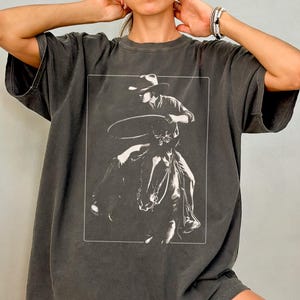 May include: A dark gray oversized t-shirt with a white graphic of a cowboy riding a horse and holding a lasso.