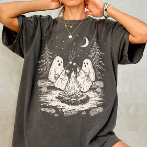 Puede incluir: Camiseta gris oscuro con un gráfico de dos fantasmas asando malvaviscos sobre una fogata en un entorno forestal. El diseño incluye una luna creciente y estrellas. La camiseta tiene mangas cortas y un corte holgado.