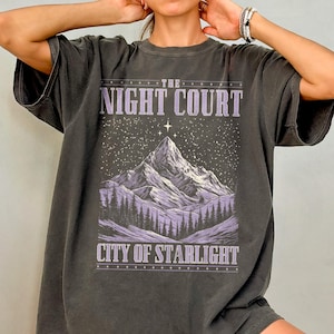 May include: A dark gray oversized t-shirt with a graphic print of a mountain range under a starry sky. The text "The Night Court City of Starlight" is printed on the shirt.