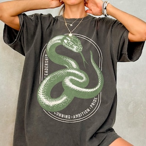 May include: Dark gray t-shirt featuring a green snake design within a white oval. The oval is encircled by the words "LEADERSHIP", "CUNNING", "AMBITION", and "PRIDE". The snake has a forked tongue.