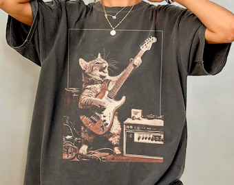May include: Dark gray oversized t-shirt featuring a cat playing an electric guitar. The cat is depicted in a vintage style, with a guitar amplifier and other musical equipment visible. The shirt has a relaxed fit and short sleeves.