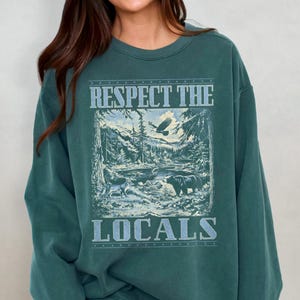 May include: A teal green sweatshirt with a graphic print of a forest scene with a bear, a wolf, and an eagle. The text "Respect the Locals" is printed in white on the sweatshirt.