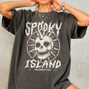 May include: Charcoal grey t-shirt with a distressed graphic of a skull with a horn, set within a sunburst design. The text reads "Spooky Island" and "Amusement Park 2002". The shirt has short sleeves and a loose fit.