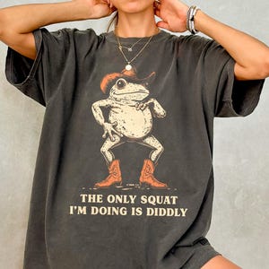 May include: Dark grey t-shirt featuring a cartoon frog wearing a cowboy hat and boots. The frog is squatting with its arms at its sides. The text "THE ONLY SQUAT I'M DOING IS DIDDLY" is printed below the frog.