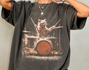 May include: Dark gray t-shirt featuring a cat playing drums. The graphic shows a cat with drumsticks, surrounded by a drum set. The shirt has a vintage, worn look and a relaxed fit.