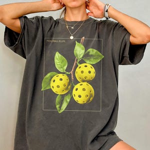 May include: Dark gray t-shirt featuring a graphic of three yellow pickleballs with green leaves and the text "Pickleball is Life" inside a white rectangle. The shirt has short sleeves and a relaxed fit.