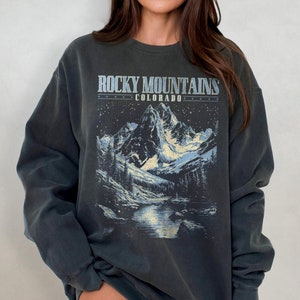 May include: A dark gray sweatshirt with a graphic print of a mountain range with a river running through it. The text "Rocky Mountains Colorado" is printed above the graphic.