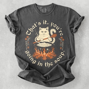 May include: Dark grey t-shirt with a humorous graphic of a cat cooking soup over a fire. The text reads "That's it, you're going in the soup." The design is surrounded by floral elements.