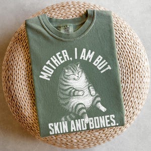 May include: Olive green t-shirt with the text "MOTHER, I AM BUT SKIN AND BONES" and a graphic of a plump cat. The shirt is made of a soft material and is laid on a woven, circular surface.