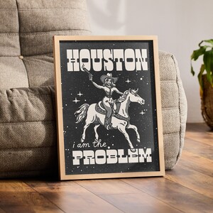 May include: A framed print with a black background featuring a woman riding a horse. The woman is wearing a cowboy hat and holding a gun. The text "HOUSTON I AM THE PROBLEM" is printed in white on the print.