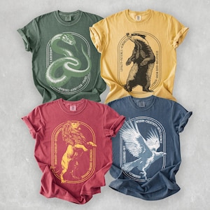 May include: Four graphic t-shirts in green, yellow, red, and blue. Each shirt features a different animal illustration inside an oval frame with text. The designs include a snake, badger, lion, and eagle, with related words.