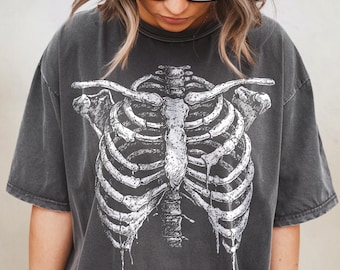 以下が含まれることがあります： 人間の肋骨の白いイラストが描かれた濃い灰色のTシャツ。グラフィックはTシャツの前面中央に配置されています。Tシャツは半袖とクルーネックです。生地は柔らかく、少し色あせたように見えます。