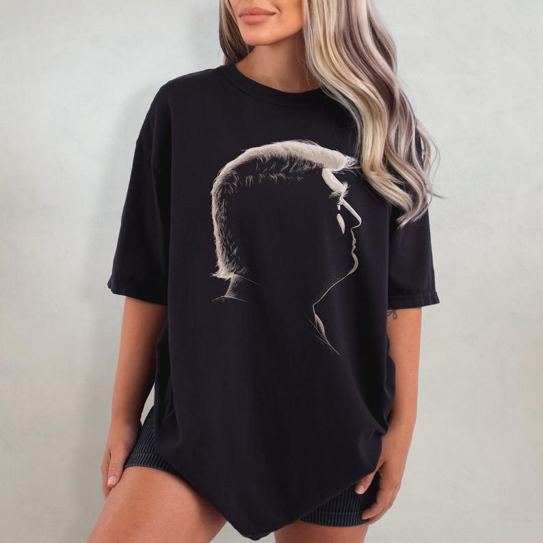 K&ouml;nnte beinhalten: Schwarzes T-Shirt mit einer Seitenprofil-Silhouette einer Person mit hellem Haar. Die Grafik ist in einer kontrastierenden hellen Farbe auf dem dunklen Stoff gehalten. Das Shirt hat kurze &Auml;rmel und eine lockere Passform.